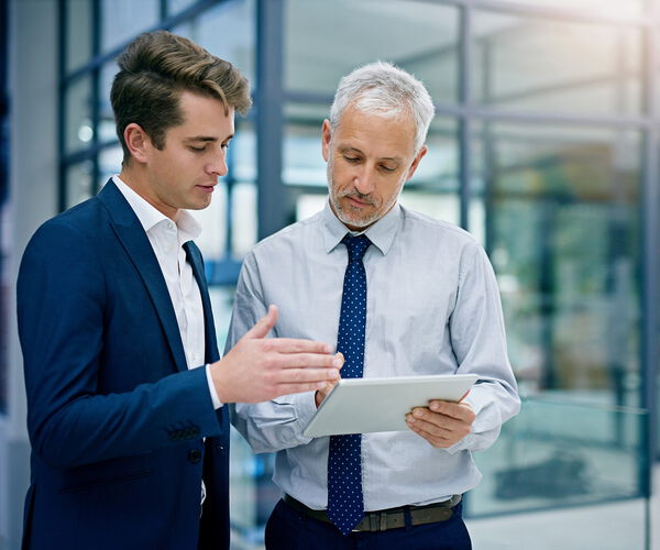 Zwei Geschäftsmänner im Gespräch in einem modernen Bürogebäude, einer zeigt dem anderen etwas auf einem Tablet.
